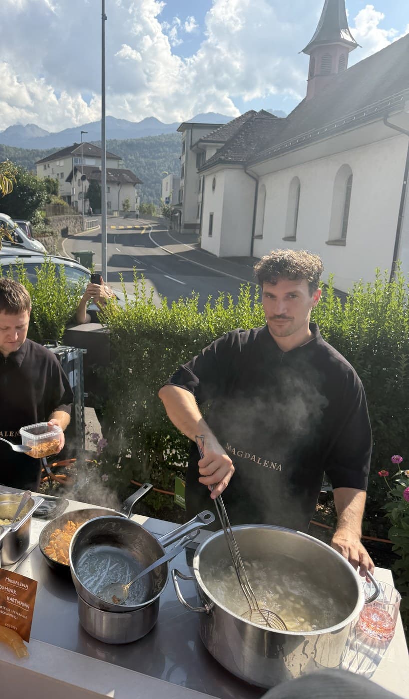 Two chefs in Magdalena polo shirts cooking cavatelli at an outdoor station, the chapel behind them
